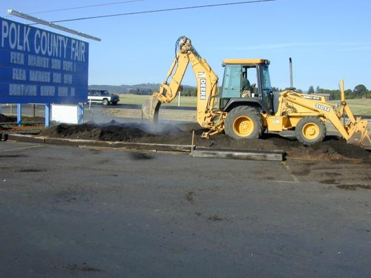 Backhoe mixing compost into compacted soil near parking lot